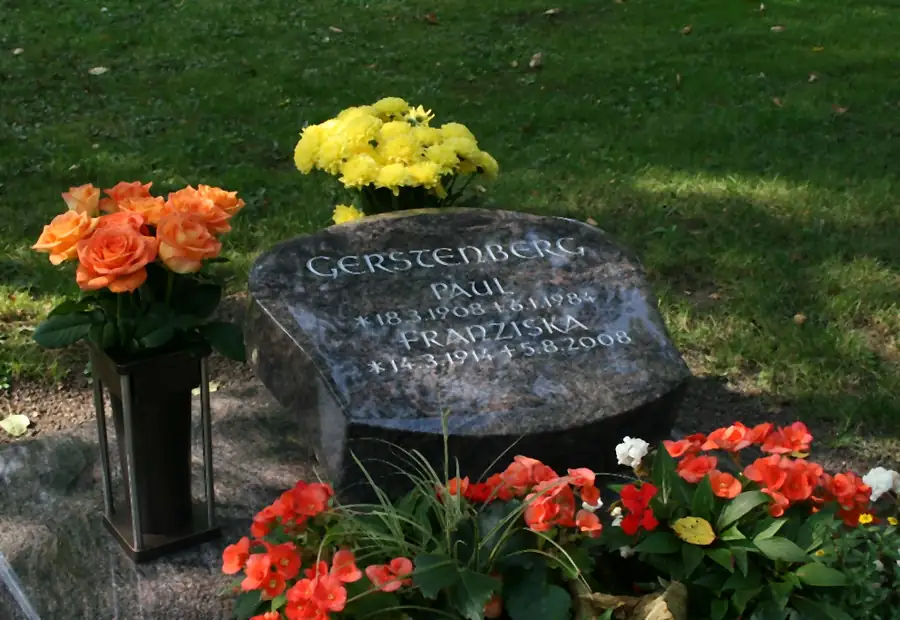 Flacher Findling als Grabmal mit eingehauener Schrift, umgeben von Bepflanzung